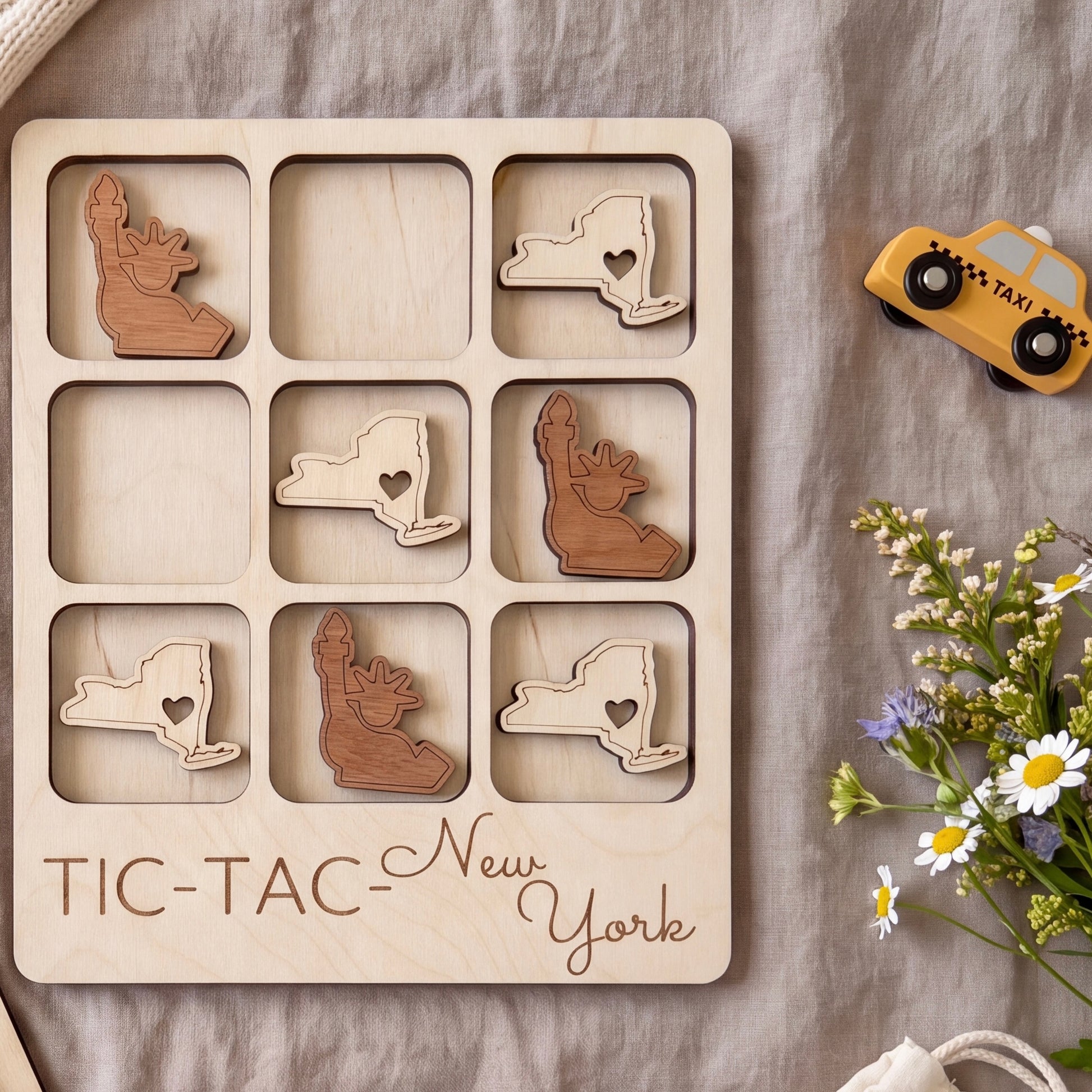 A wooden tic-tac board with various shapes cut out, including a heart-shaped piece, a wooden bear, and a wooden car, along with a toy taxi and some flowers.