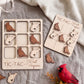 a wooden tic-tac game with a bird-shaped tile and a wooden tic-tac card with a bird-shaped tile, both placed on a grey surface. There is also a red bird figurine and some leaves in the background.
