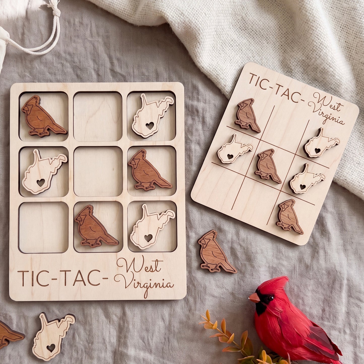 a wooden tic-tac game with a bird-shaped tile and a wooden tic-tac card with a bird-shaped tile, both placed on a grey surface. There is also a red bird figurine and some leaves in the background.