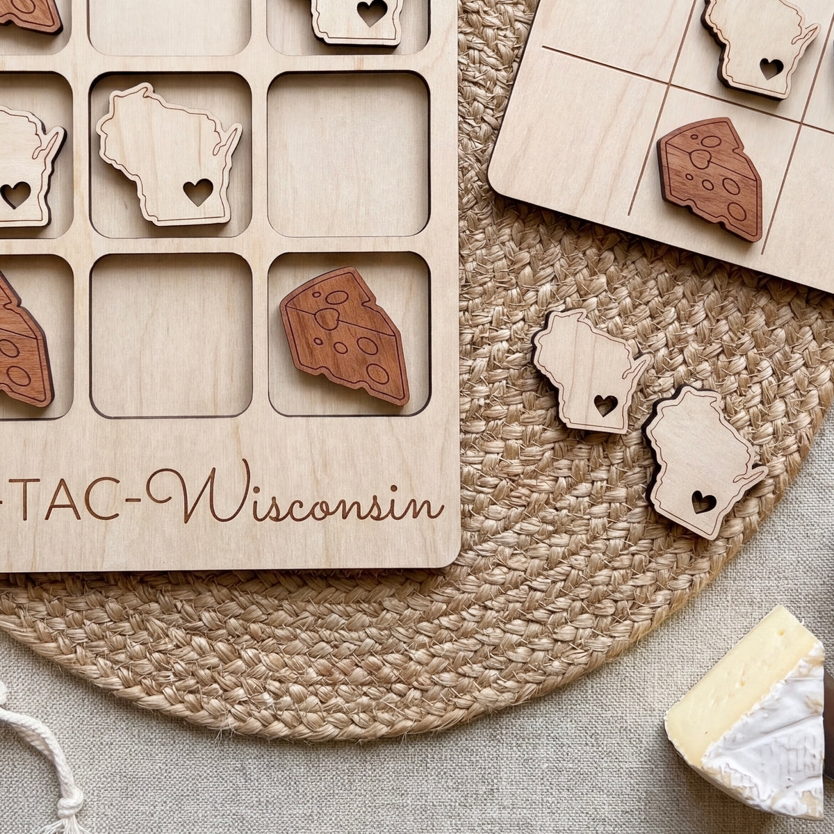 a wooden game board with a Wisconsin theme, along with several wooden pieces shaped like cheese and hearts. The game board is placed on a woven placemat, and there is a piece of cheese visible in the bottom right corner of the image.
