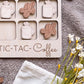 a wooden tic-tac coffee game board with coffee mug and bean shapes as game pieces, placed on a gray surface alongside a white cloth and some flowers.