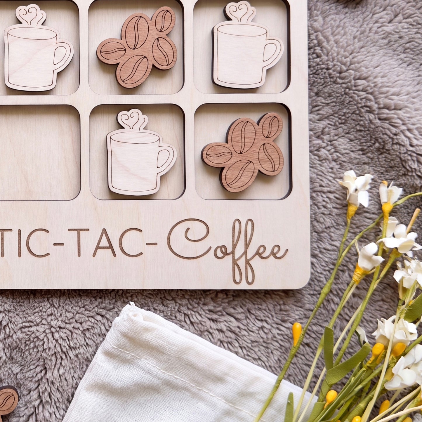 a wooden tic-tac coffee game board with coffee mug and bean shapes as game pieces, placed on a gray surface alongside a white cloth and some flowers.