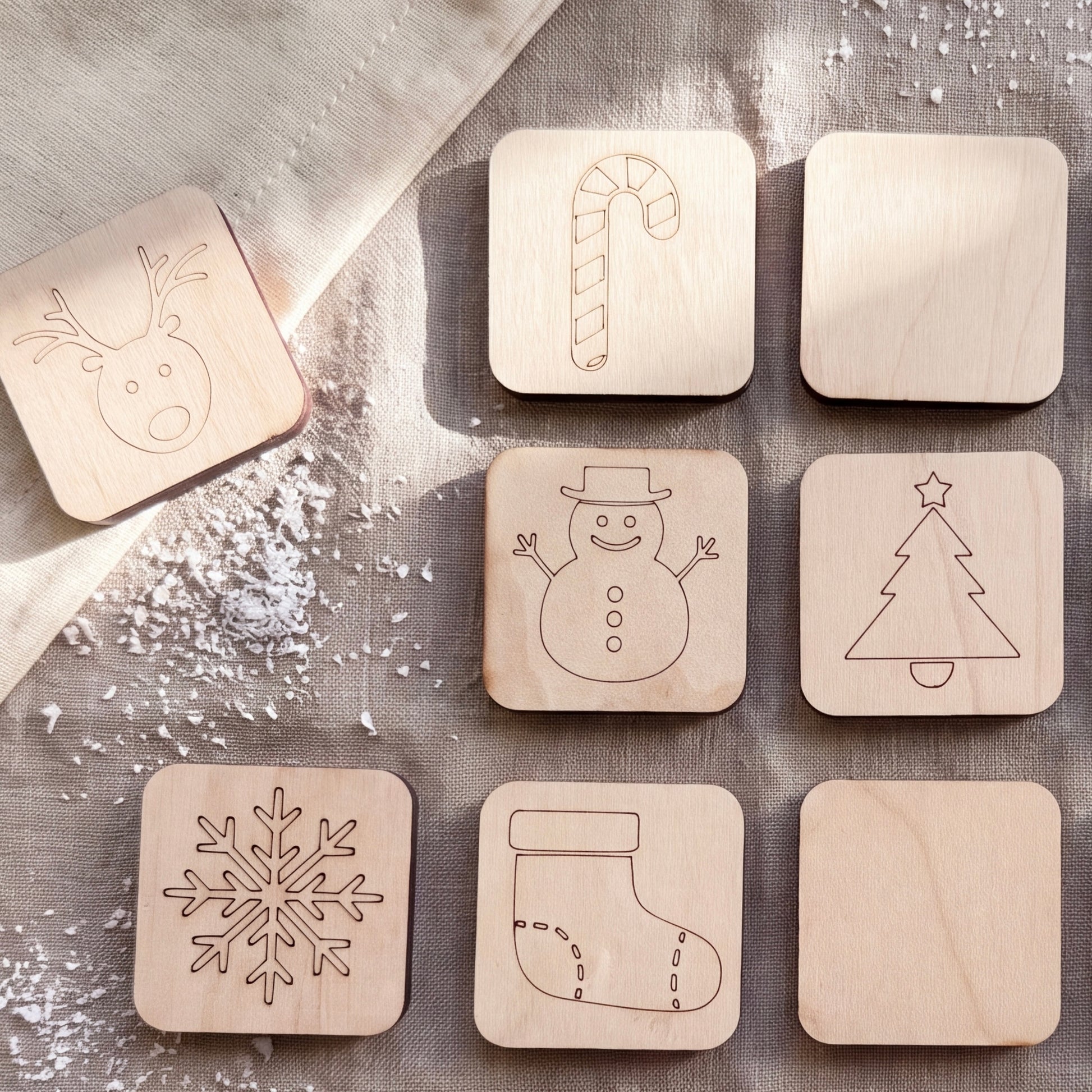 A collection of wooden coaster-shaped objects, each featuring a different winter-themed design, including a reindeer, candy cane, snowman, snowflake, and stocking.