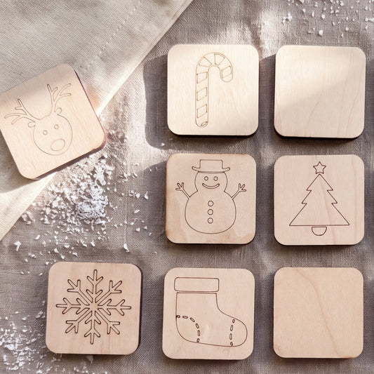 A collection of wooden coaster-shaped objects, each featuring a different winter-themed design, including a reindeer, candy cane, snowman, snowflake, and stocking.