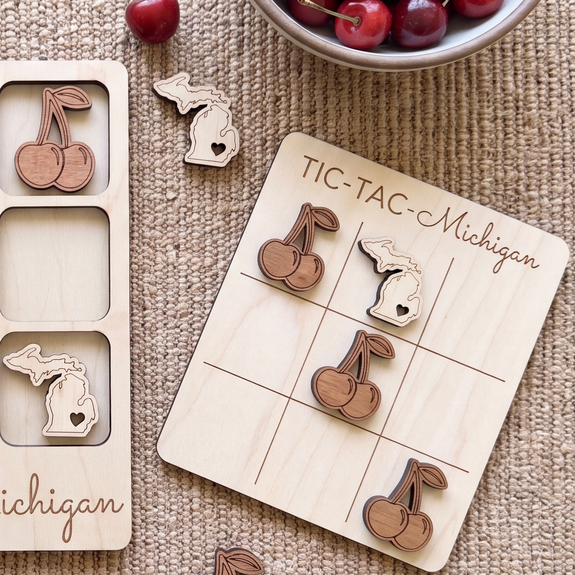 a wooden tic-tac game board with wooden cherries and a bowl of cherries on a woven surface.