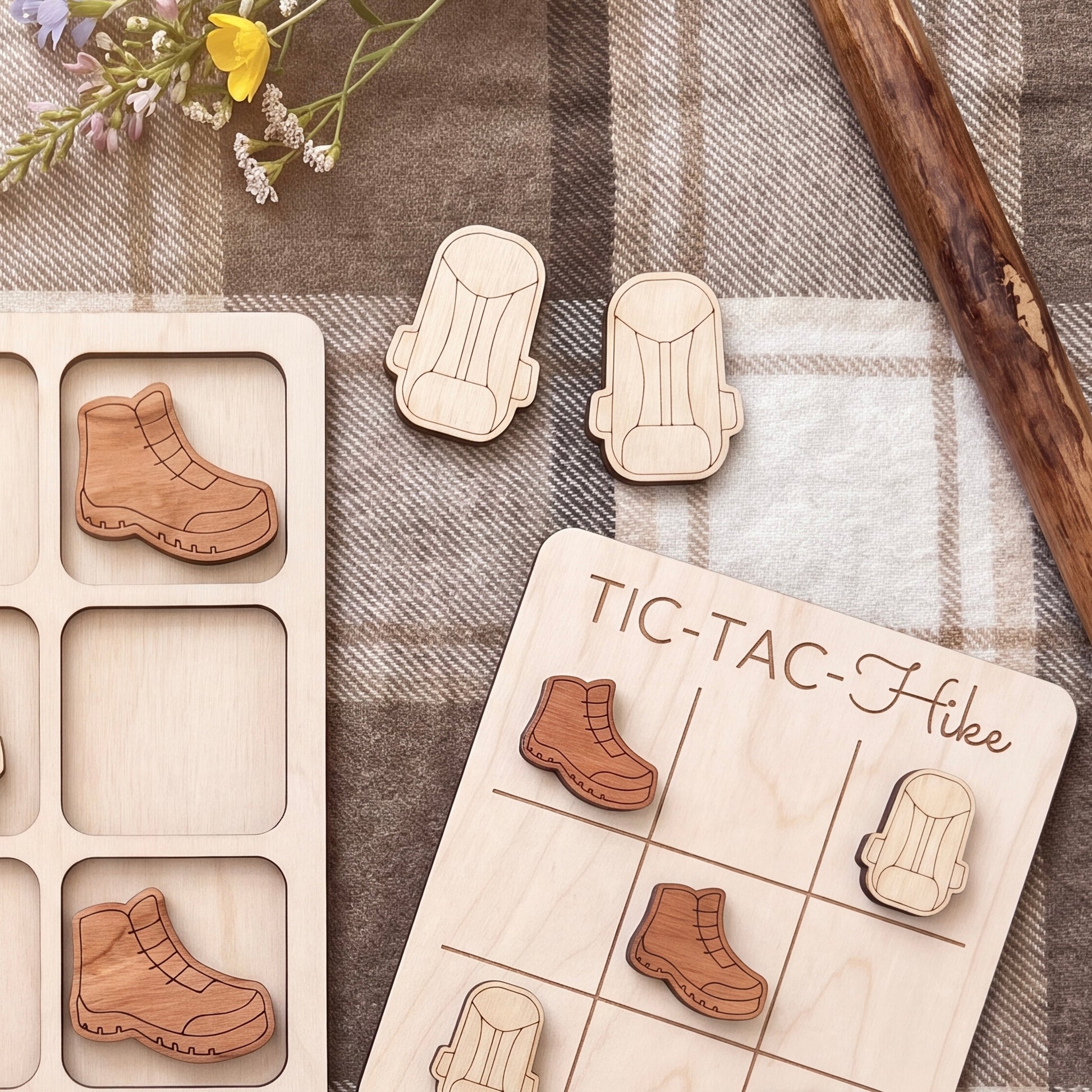 a wooden tic-tac game board with wooden pieces shaped like hiking boots, a wooden stick, and a floral arrangement on a plaid cloth.