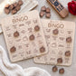 two wooden bingo cards with various TV-related questions and bingo squares, along with several wooden bingo pieces and a remote control on a beige surface.