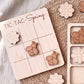 A wooden tic-tac game board labeled "TIC-TAC-Spring" with wooden tic-tac pieces featuring a bee and a flower design.