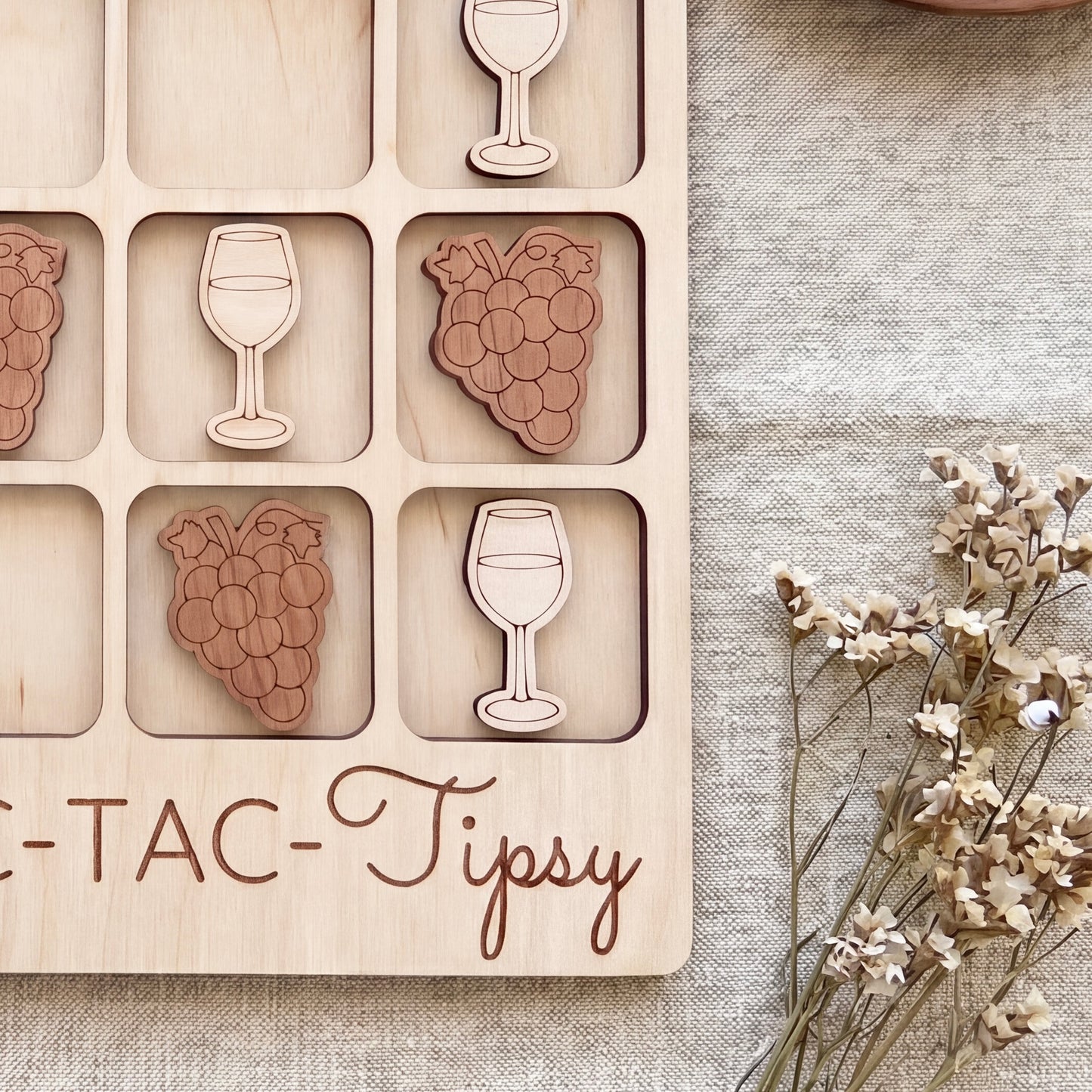 a wooden tic-tac game board with wooden pieces shaped like wine glasses and grapes, placed on a beige cloth.