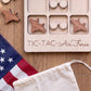 a wooden tic-tac game with a wooden airplane and a wooden pair of sunglasses on it, along with a white cloth and a red and white American flag.