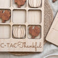 A wooden tic-tac-toe board with wooden turkey and pumpkin tiles, along with a wooden placemat with the phrase "C-TAC-Thankful" written on it.