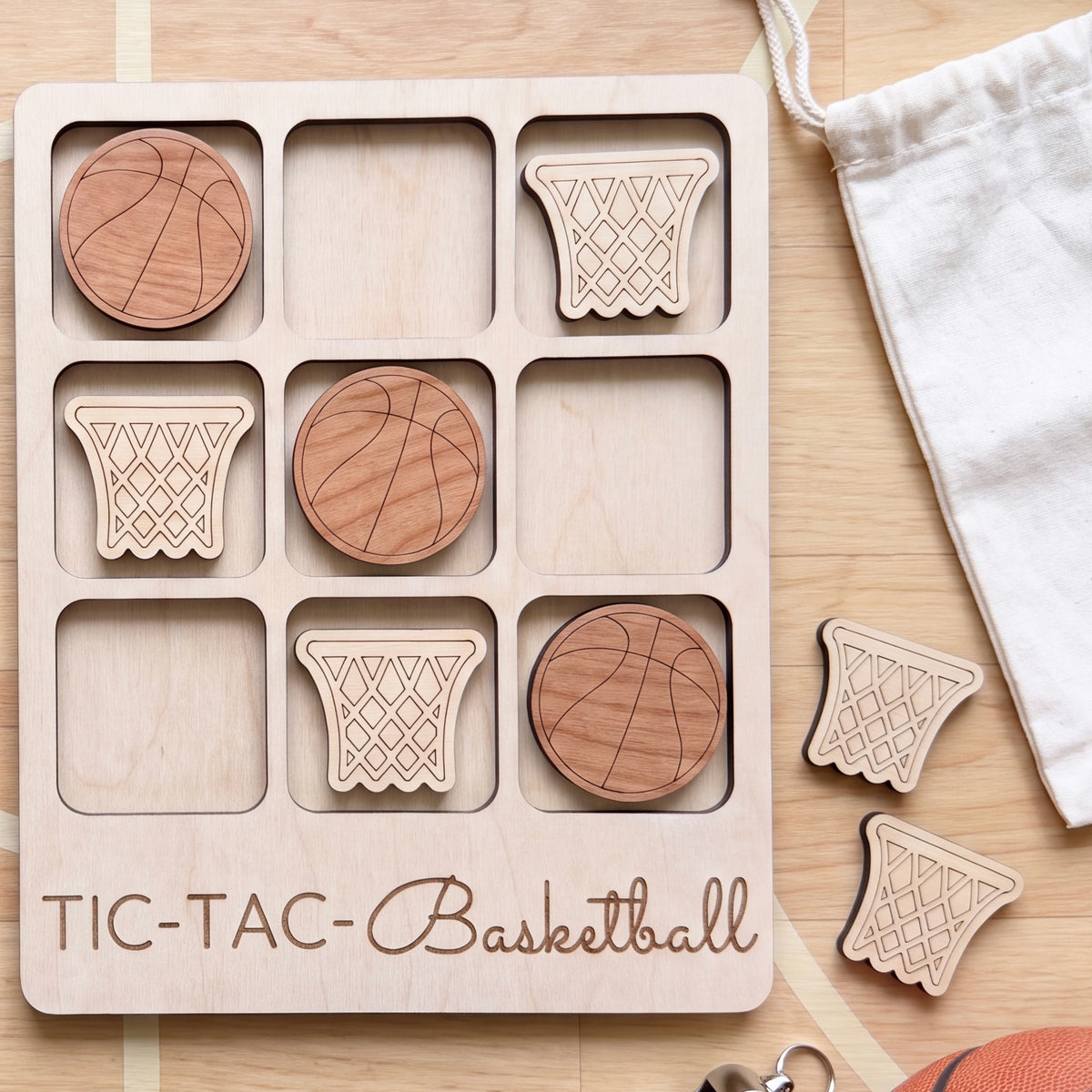 A wooden tic-tac game board with wooden basketballs and a white drawstring bag is displayed on a wooden surface.