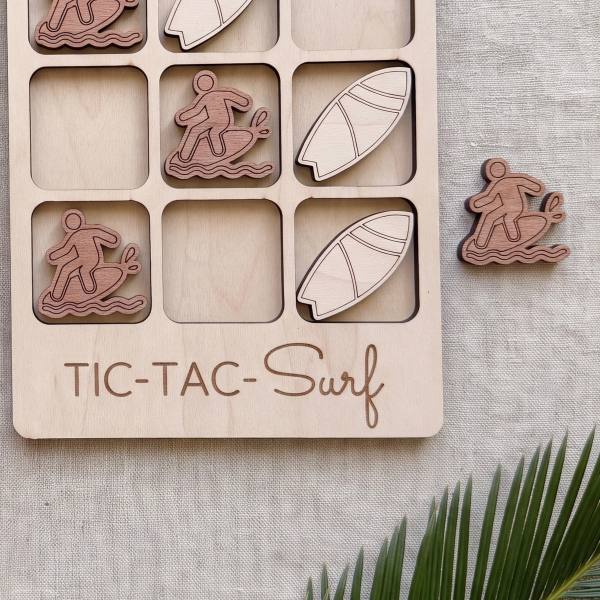 a wooden game board with a tic-tac game design, along with a small wooden surfboard and a palm frond.