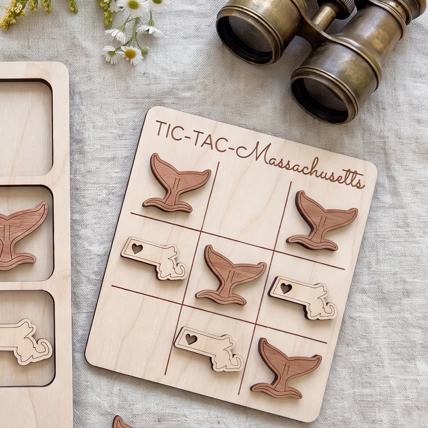 A wooden tic-tac game board with whale-shaped tiles, accompanied by a pair of binoculars and some flowers on a table.