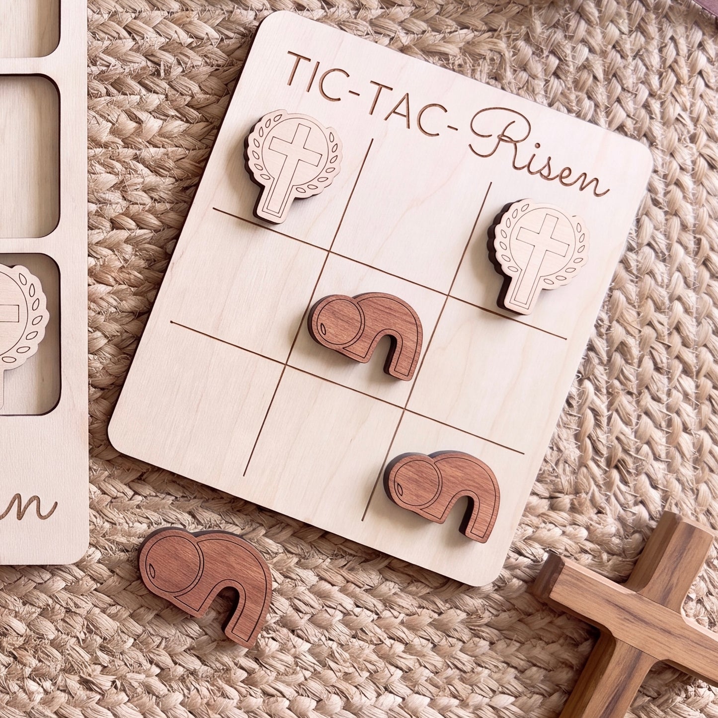 a wooden tic-tac game board with wooden pieces, including a cross and a heart, arranged on a woven surface.