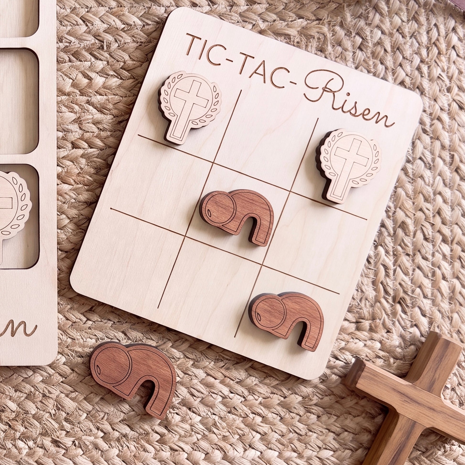 a wooden tic-tac game board with wooden pieces, including a cross and a heart, arranged on a woven surface.