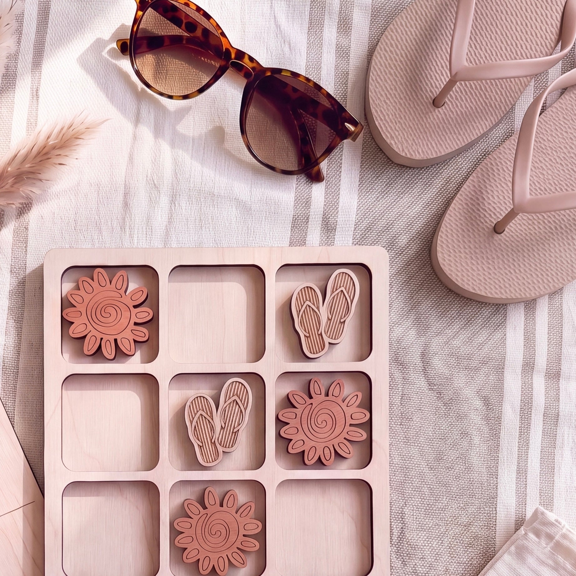 A wooden tic-tac-toe board with wooden tiles, a pair of sunglasses, and a pair of flip-flops on a striped towel.