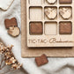 a wooden tic-tac board game with wooden pieces shaped like books and apples, placed on a beige cloth.