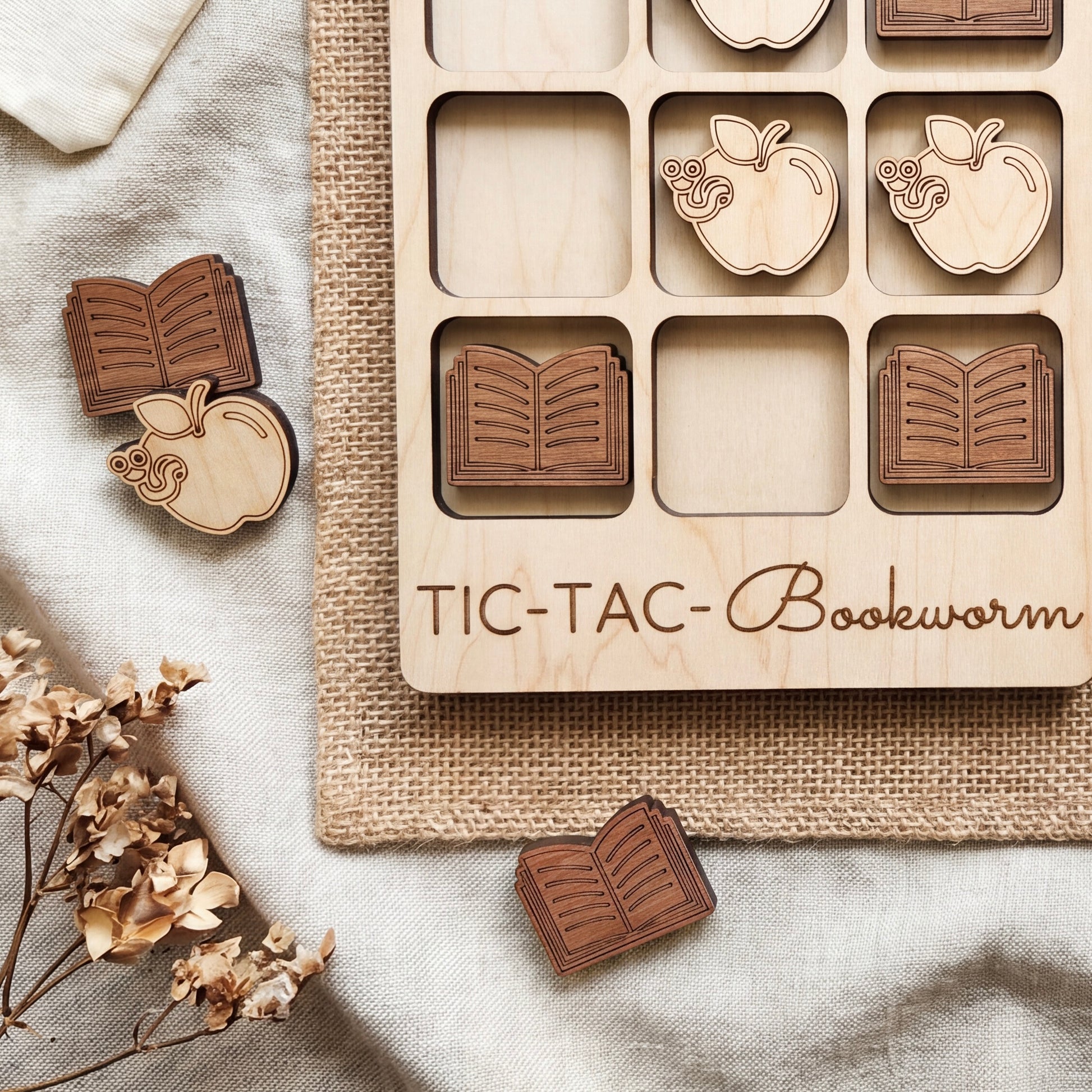 a wooden tic-tac board game with wooden pieces shaped like books and apples, placed on a beige cloth.