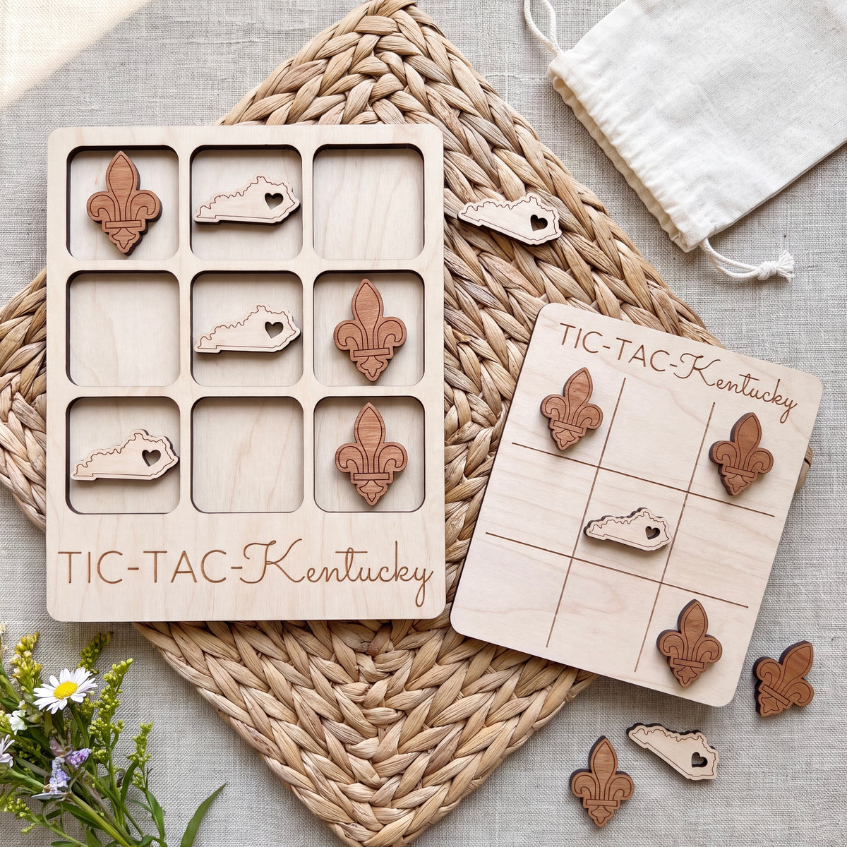 a wooden tic-tac game board and a tic-tac card with wooden pieces, both labeled "TIC-TAC-KENTUCKY". The game board and card are placed on a woven placemat, and there are some flowers visible in the background.