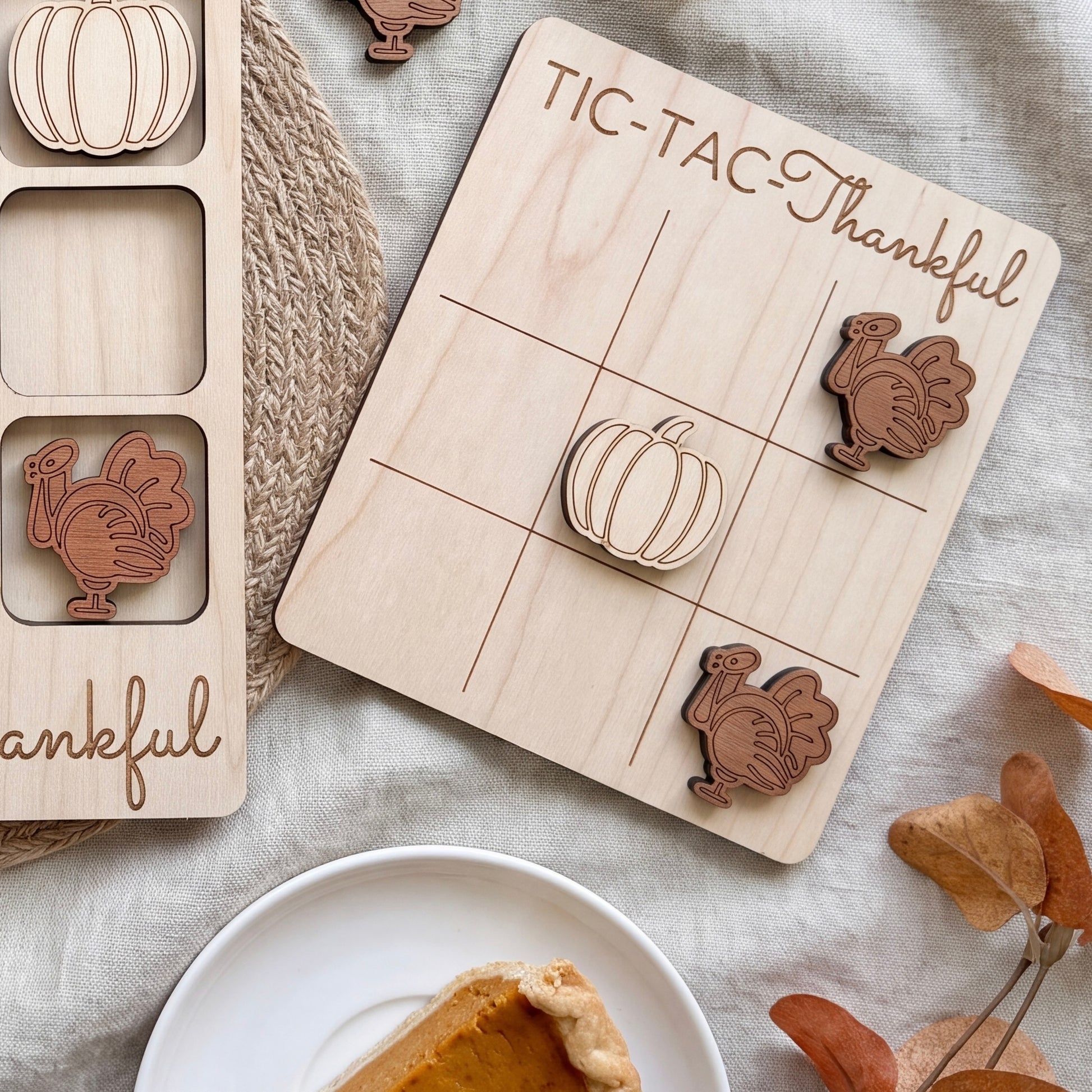 a wooden tic-tac game board with Thanksgiving-themed wooden pieces, including a turkey and a pumpkin. The game board is placed on a table, and there is a slice of pumpkin pie and some autumn leaves in the background.