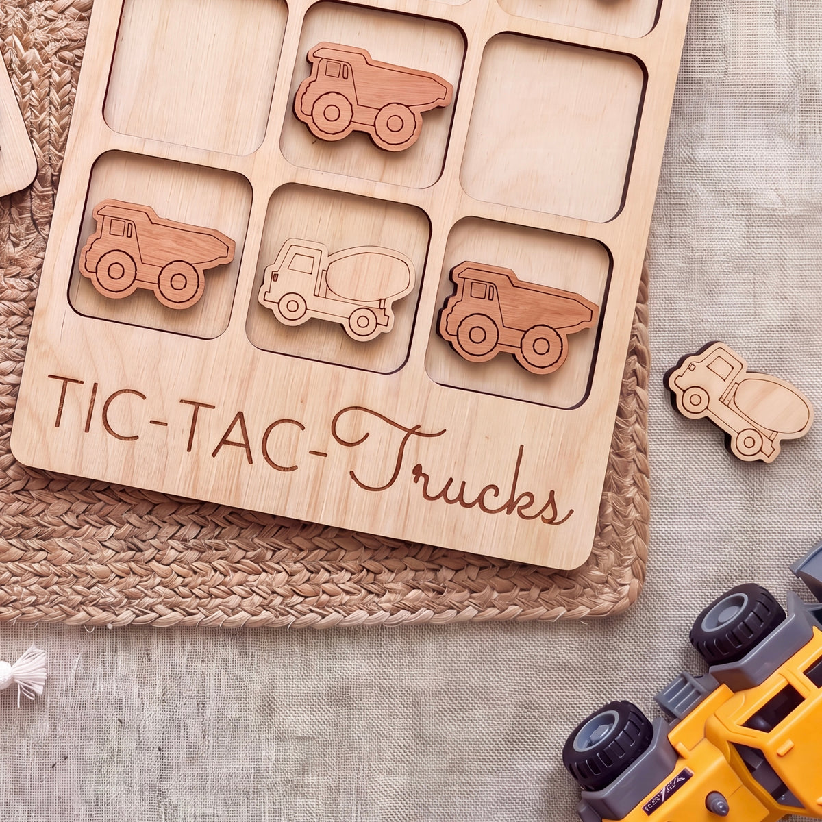 A wooden tic-tac-toe game board with wooden truck tiles on it, along with a yellow toy truck and a woven placemat.
