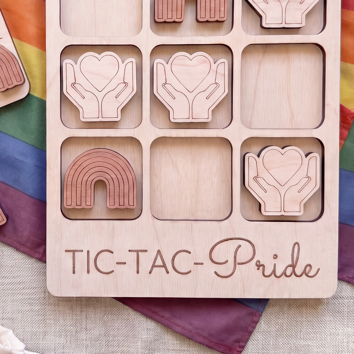 A wooden tic-tac game with the word "TIC-TAC-Pride" written on it, along with various heart-shaped and rainbow-shaped tiles.