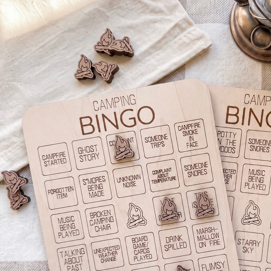 a wooden bingo board with various camping-related questions and bingo cards, along with some wooden bingo pieces on a white cloth.