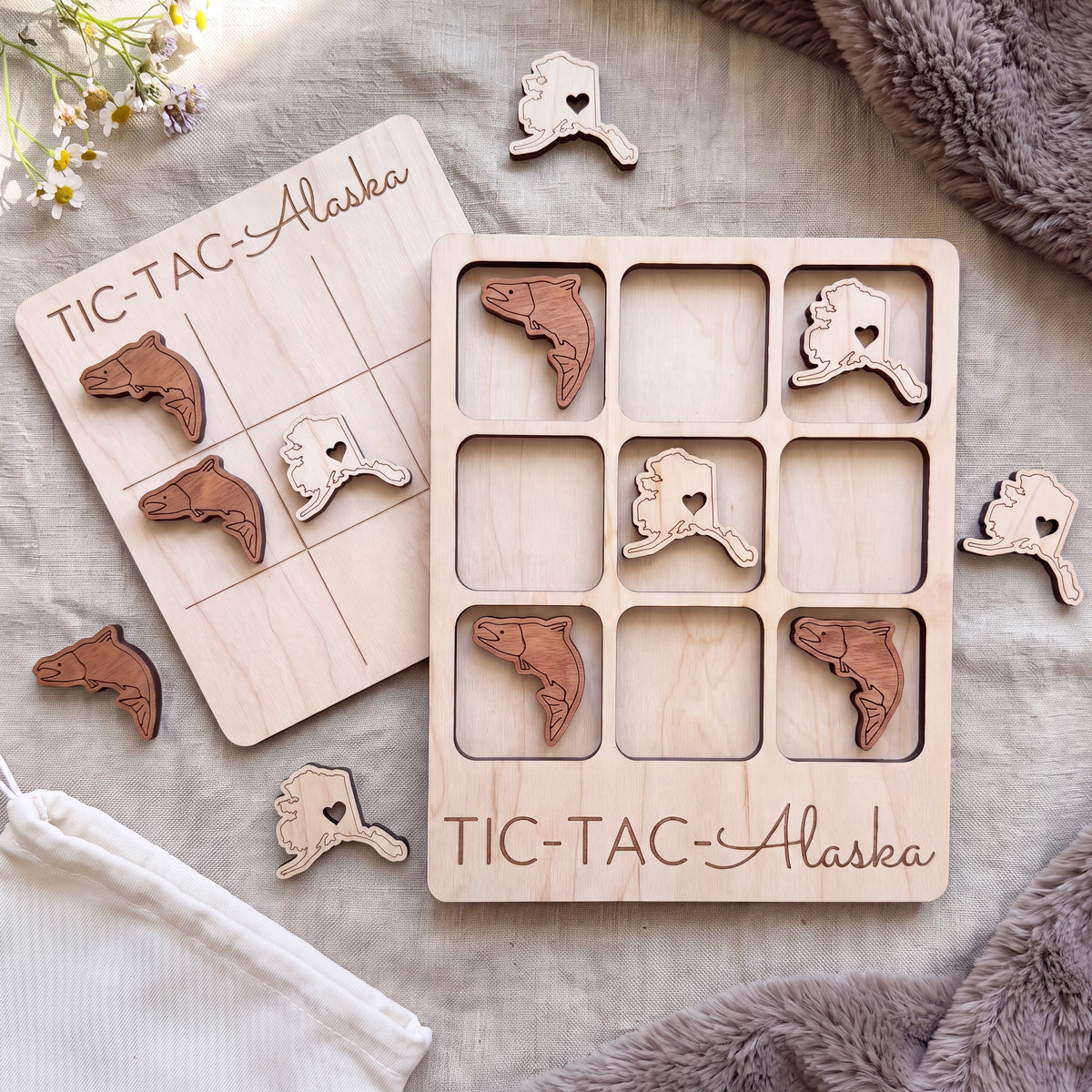 a wooden Tic-Tac-Alaska game board and several wooden pieces shaped like fish, arranged on a grey fabric surface.