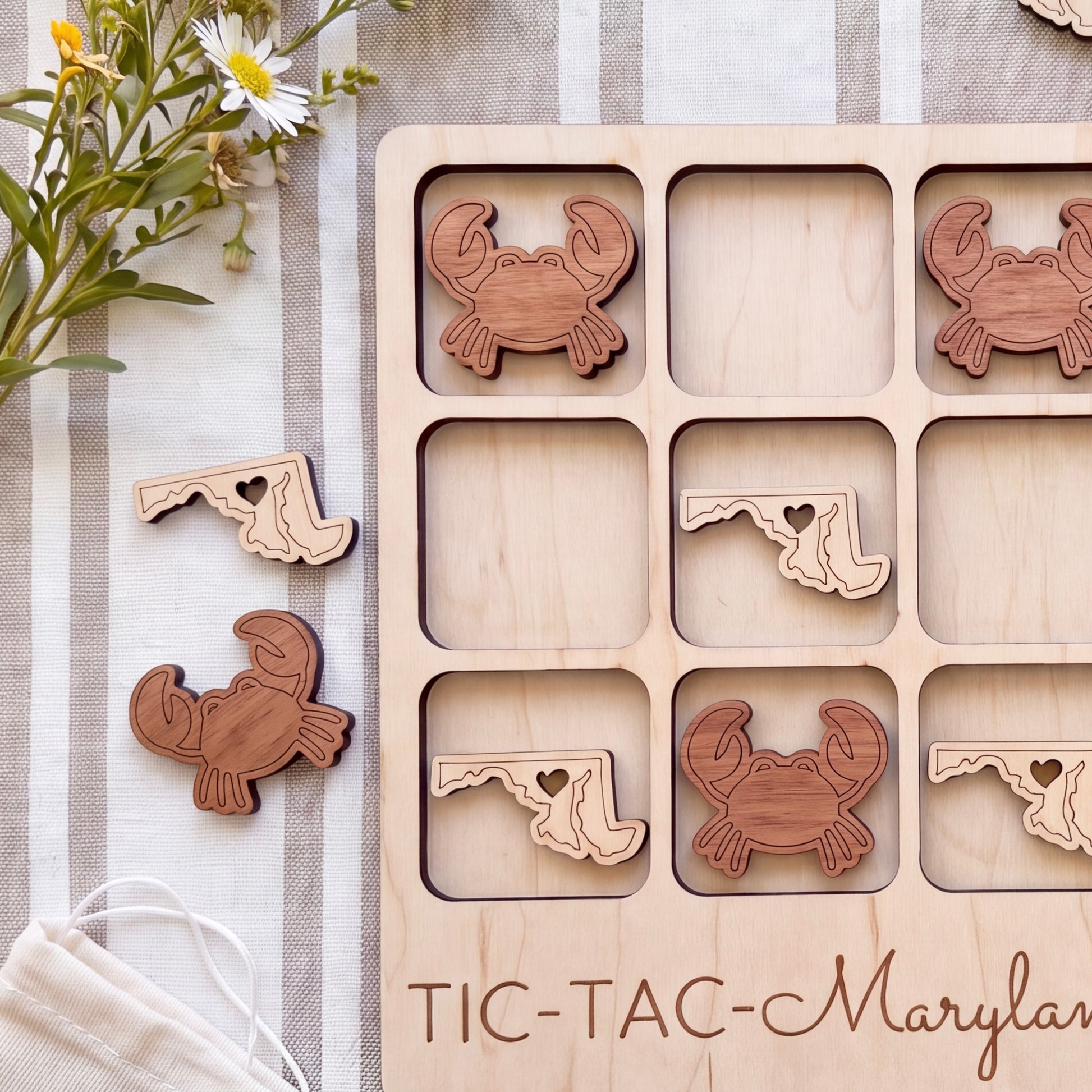A wooden tic-tac game board with wooden crab and lobster pieces, along with a bouquet of flowers and a striped cloth in the background.