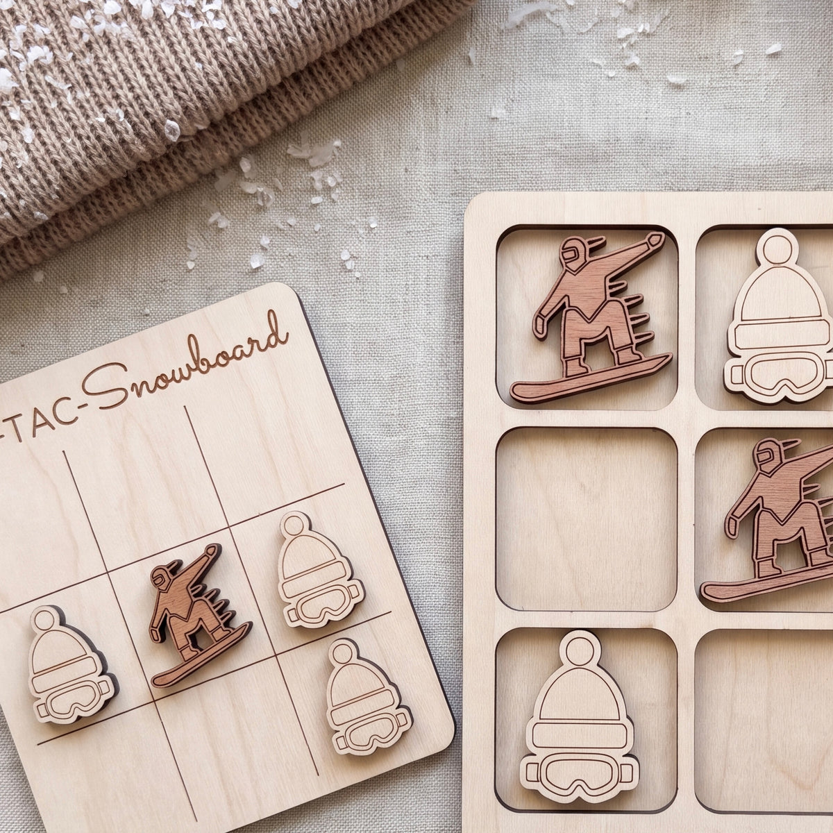 a wooden snowboard-themed game board and a wooden snowboard with various snowboarder icons, all placed on a beige fabric surface.