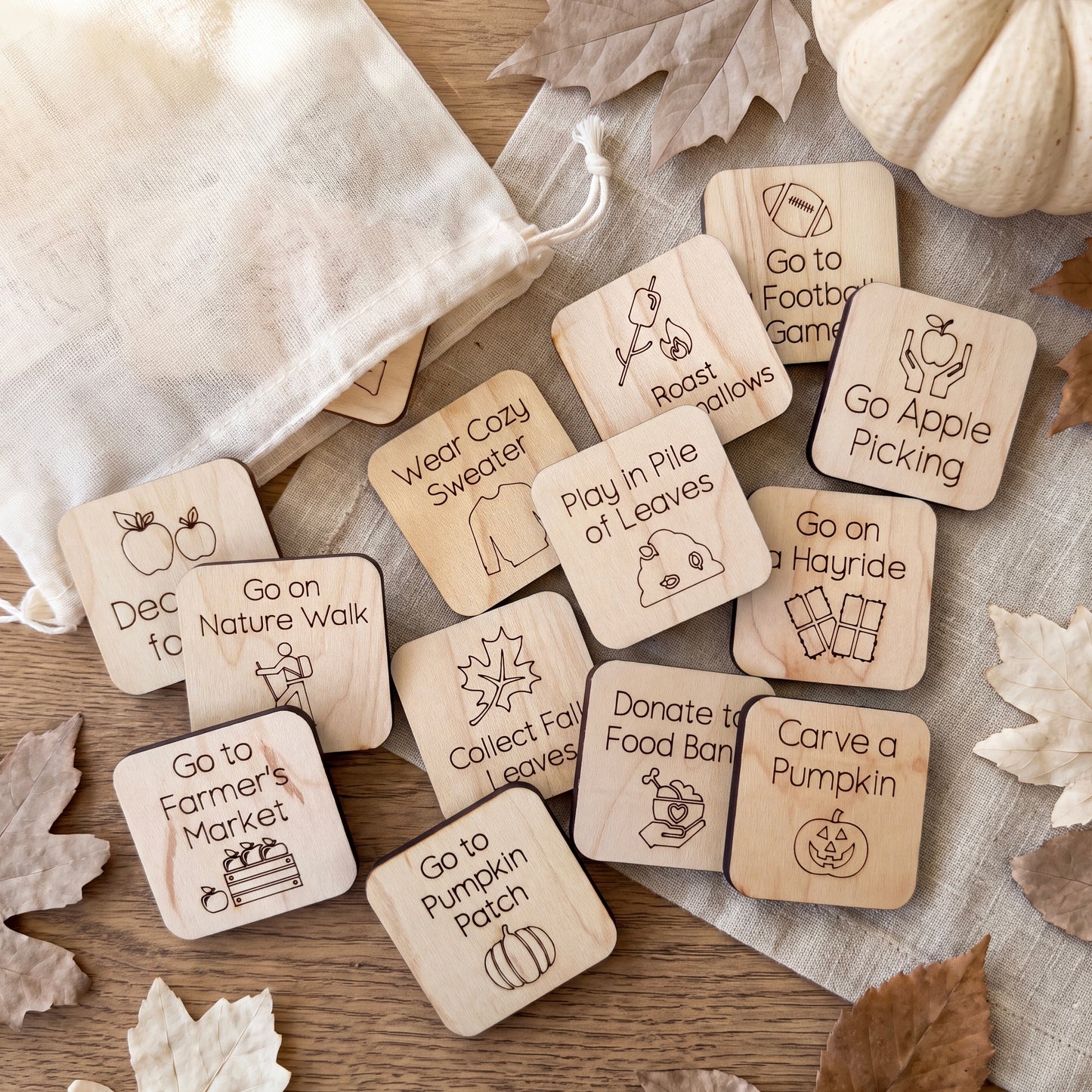 a collection of wooden tags with various fall-themed activities and instructions, such as "Go to Nature Walk", "Go to Farmer's Market", "Go to Wear Cozy Sweater", "Go to Play in Pile of Leaves", "Go on Hayride", "Donate to Food Bank", "Carve a Pumpkin Patch", and "Go on Nature Walk". The tags are scattered on a wooden surface, with some leaves visible in the background.