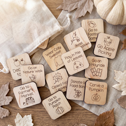 a collection of wooden tags with various fall-themed activities and instructions, such as "Go to Nature Walk", "Go to Farmer's Market", "Go to Wear Cozy Sweater", "Go to Play in Pile of Leaves", "Go on Hayride", "Donate to Food Bank", "Carve a Pumpkin Patch", and "Go on Nature Walk". The tags are scattered on a wooden surface, with some leaves visible in the background.