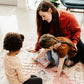 A woman and two young girls are playing a game on a patterned rug in a cozy living room.