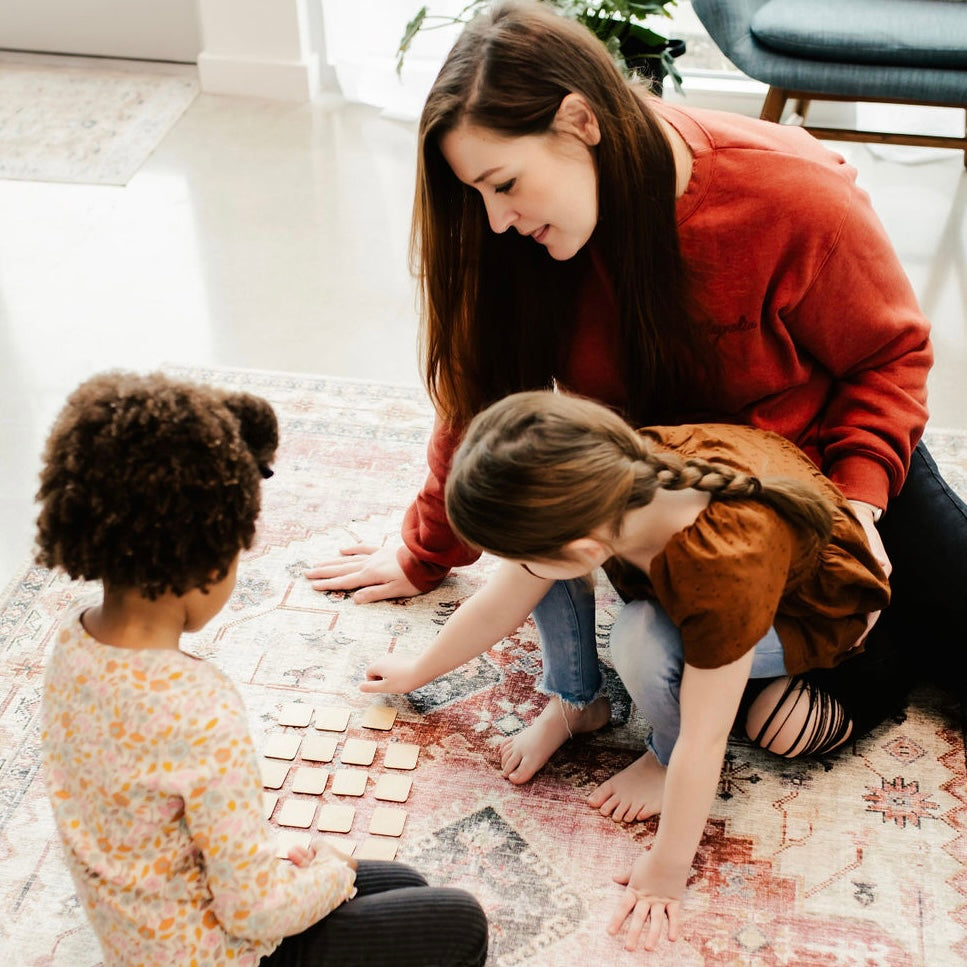 A woman and two young girls are playing a game on a patterned rug in a cozy living room.